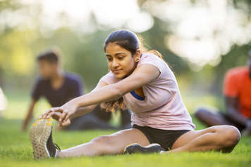 Teen sitting in grass with one leg stretched out while bending forward to touch her toes on that foot with both hands