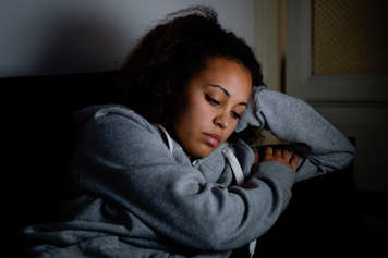 Teen girl sitting on a couch in the dark, leaning to the side to rest her head on one arm that rests on the couch arm
