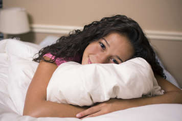 Teen girl lying on her stomach in bed and hugging a pillow under her head while smiling at the camera