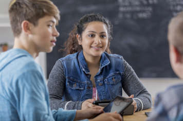 Group of 3 teens sitting around a desk in a classroom and holding tablets. Two of the teens look at the third teen